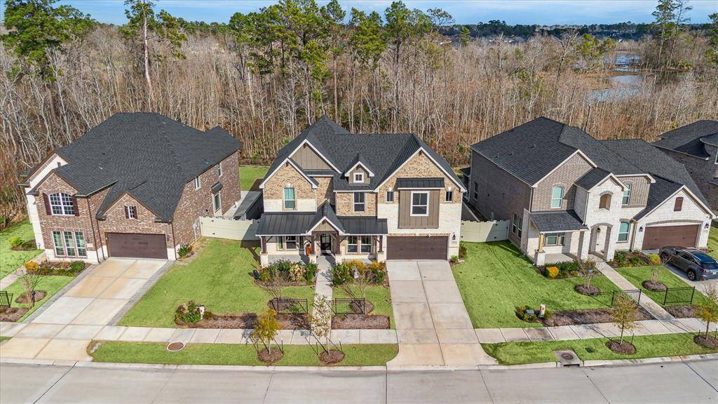 This aerial image of the house showcases a suburban neighborhood and is a modern two-story home with a combination of brick and stone exterior, and a covered front porch. It features a two-car garage, a well-manicured lawn with landscaping, and a fenced backyard, all set against a wooded backdrop.