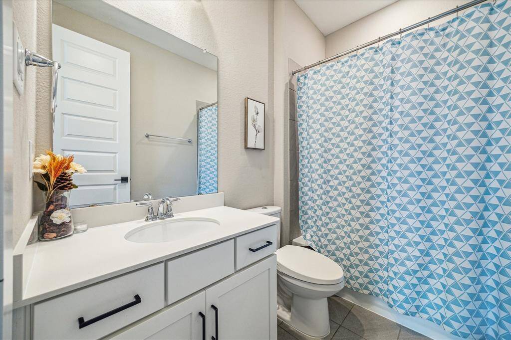 This is the second bathroom (attched to 2nd bedroom) on the first floor, featuring a white vanity with a sleek countertop and black hardware. The space is complemented by a modern blue geometric shower curtain and a large mirror that enhances the brightness of the room.
