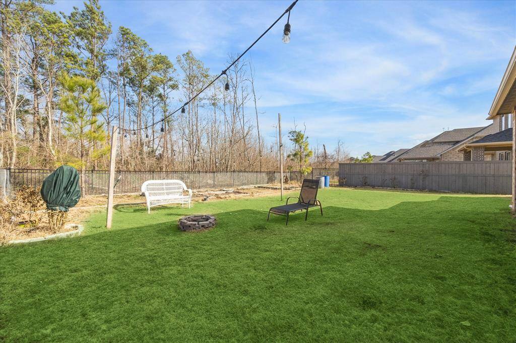 The backyard features a spacious lawn with a designated flower bed along the fence line, adding a natural touch to the outdoor space. A fire pit sits in the center, creating a cozy gathering area beneath overhead string lights.