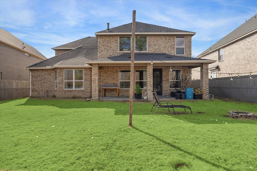 The back view of the house showcases a brick exterior with a covered patio extending into the spacious backyard. String lights are hung across the yard, enhancing the inviting outdoor atmosphere.