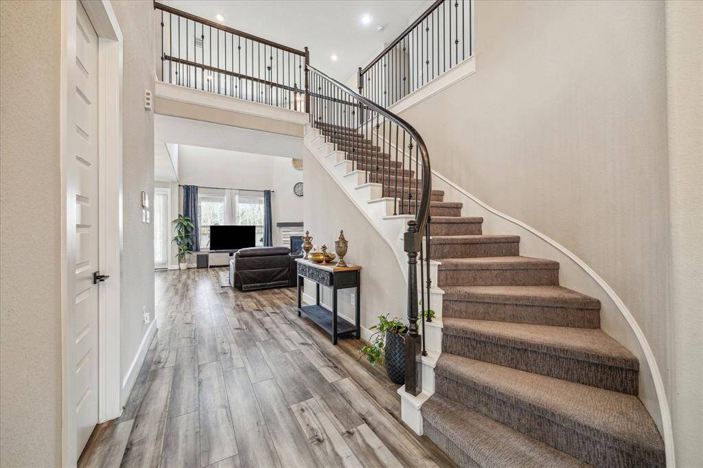 a grand entryway with a curved staircase featuring elegant wrought-iron railings and carpeted steps. The open-concept design leads to a spacious living area with high ceilings, large windows, and a modern fireplace, creating a bright and inviting atmosphere.