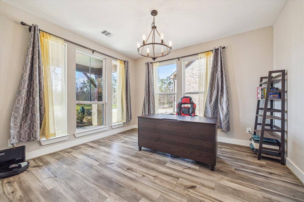This bright and inviting home office features large windows that fill the space with natural light and offer scenic outdoor views. The room is enhanced by wood-look vinyl flooring, a stylish chandelier, and a modern setup ideal for productivity.