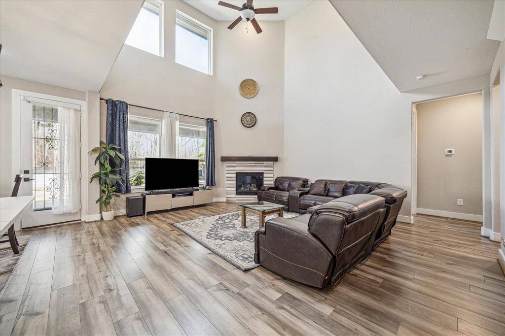 This spacious living room features soaring ceilings, large windows that bring in abundant natural light, and a modern fireplace as a focal point. The open layout is complemented by sleek wood-look vinyl flooring.