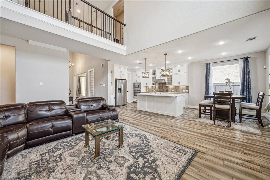This open-concept space seamlessly connects the living area to a modern kitchen featuring white cabinetry, a large island, and stylish pendant lighting. Natural light flows in through large windows, highlighting the elegant flooring and spacious layout.
