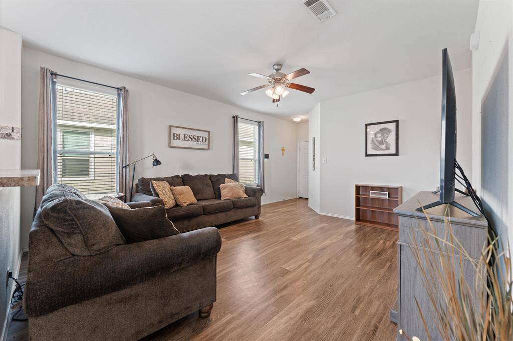 Bright and open living area with luxury vinyl flooring and neutral walls and large windows letting in natural light. The space flows into a modern kitchen with a breakfast bar, stainless steel appliances, and ample cabinetry.