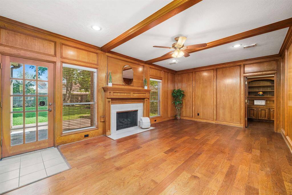 Another view of the spacious family room accents warm wood paneling, cozy fireplace, and ceiling beams. Natural light pours in through large windows and a glass door that opens to the serene backyard.