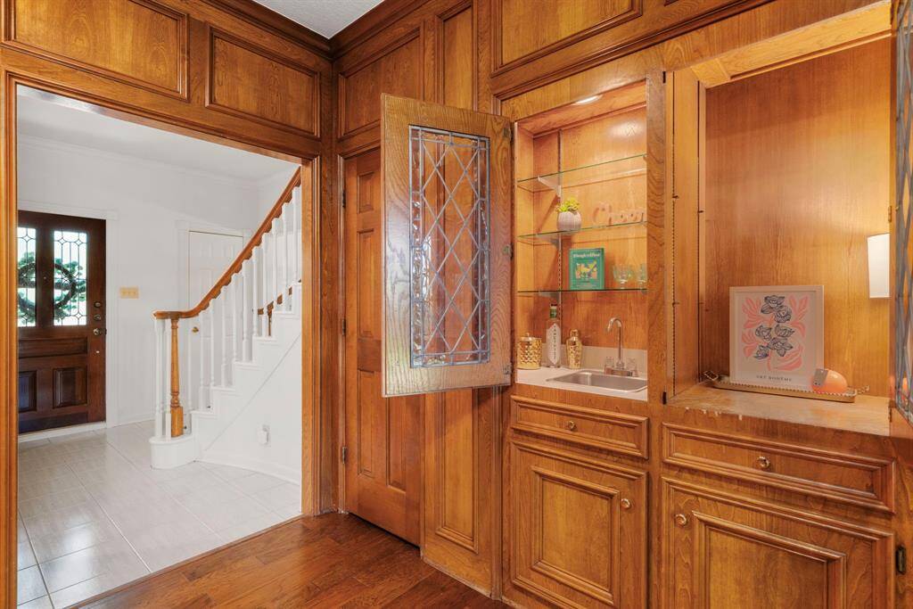 This family room photo showcases the elegant wood paneling, built-in wet bar area with glass shelving and a small sink. The space is warm, inviting, and perfect for entertaining & welcoming guests.