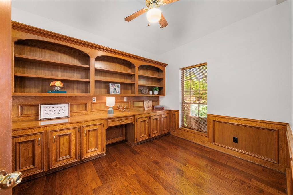 This view of the cozy home office shows the ceiling fan, a large window that provides natural light, & a view of the front landscape. The warm wood tones create an inviting atmosphere.