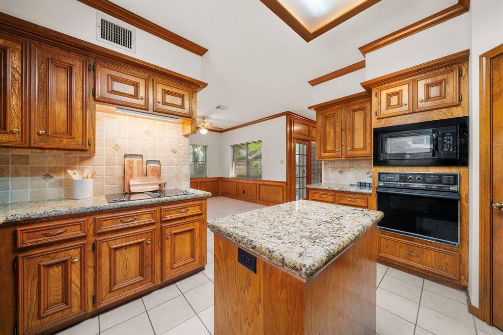 This kitchen features warm wood cabinetry, granite countertops, and tile backsplash. It includes a central island, a built-in microwave, and a wall oven. Ample natural light enhances the open layout, with a view into the large sunny breakfast room.