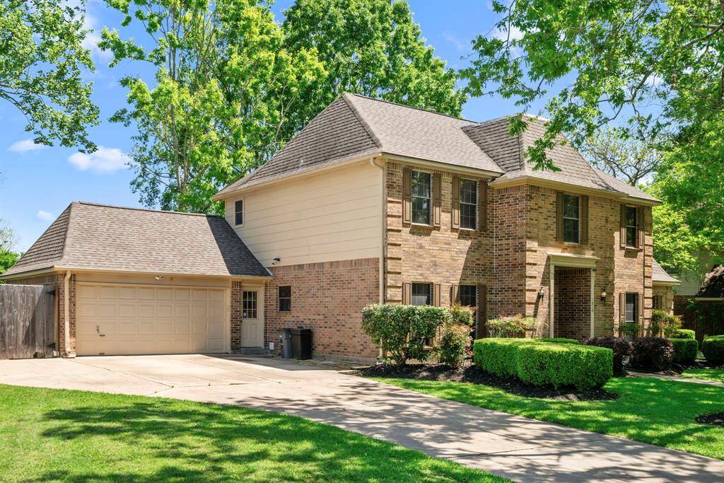 This is a two-story brick home with an attached two-car, attached garage, extra parking space, and well-maintained landscaping. The property is surrounded by mature trees, providing a pleasant, shaded environment.