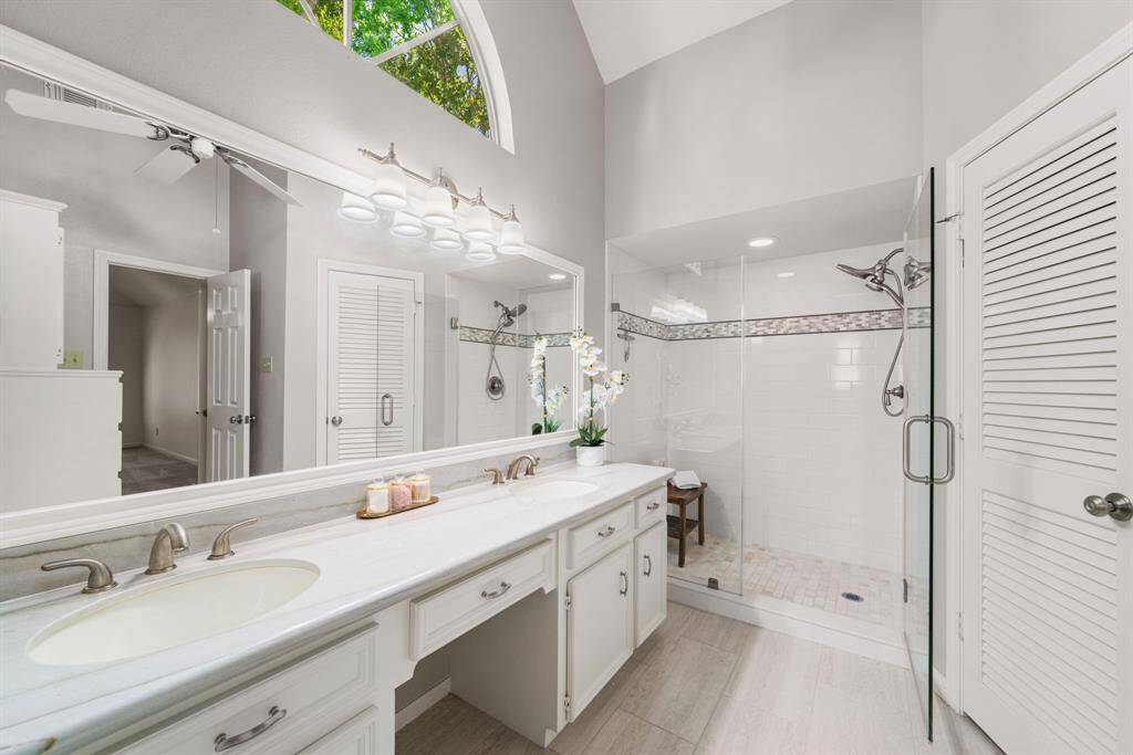 This Primarybathroom features a spacious, modern design with dual sinks, a large mirror, and ample lighting. It includes a glass-enclosed shower with elegant tile detailing, a vaulted ceiling, and neutral tones for a fresh, airy feel.