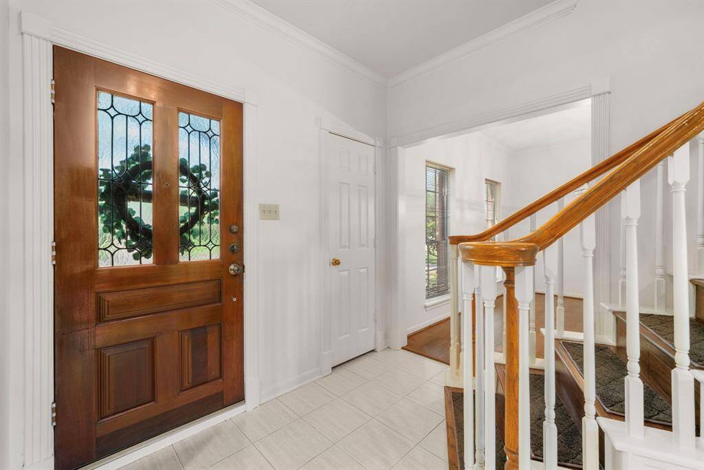 A welcoming entryway featuring a wooden door with decorative glass panels, bright white walls, and a staircase with wooden handrails. Natural light streams in from nearby windows, creating an inviting atmosphere.