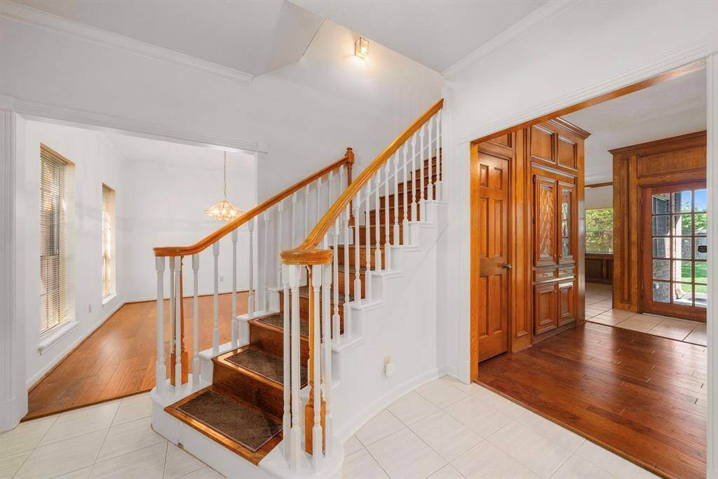 This welcoming entryway features a bright, open space with a wooden staircase, tile flooring, and warm wood accents. Large windows allow natural light, complemented by elegant light fixtures. The space leads to adjoining rooms with rich wood details and views of the outdoors.