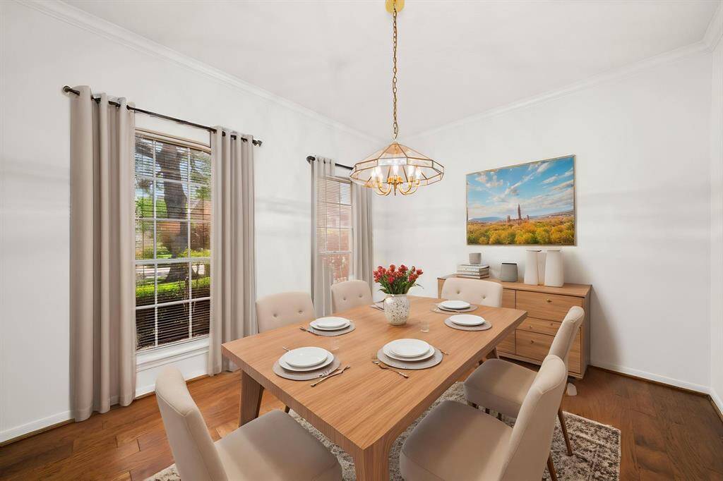 Bright dining room with large windows, neutral walls, and wood floors. Features a modern hanging light fixture, and is virtually staged with a wooden table set for six, and a buffet with decor.