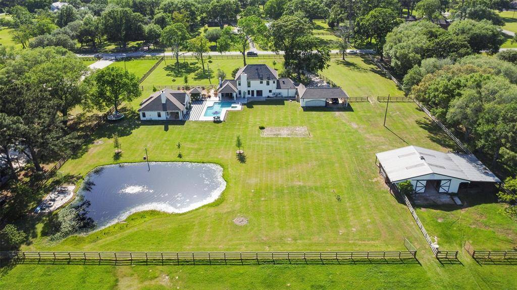 Aerial view of the property from the cross fence depicting the back of the barn, pond, firepit, guest house, pool, main house and detached garage and carport.  Note the lush mature trees that provide total privacy on either side.
