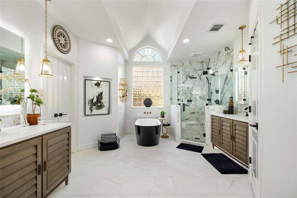 A private sanctuary, reimagined. Completely remodeled in 2022, this luxurious primary bathroom combines modern sophistication with timeless design. The expansive layout features dual vanities, sleek black and white finishes, and elegant touches throughout. A vaulted ceiling and large glass block window bathe the space in natural light, while upgraded fixtures and custom details elevate the everyday experience to something truly exceptional.