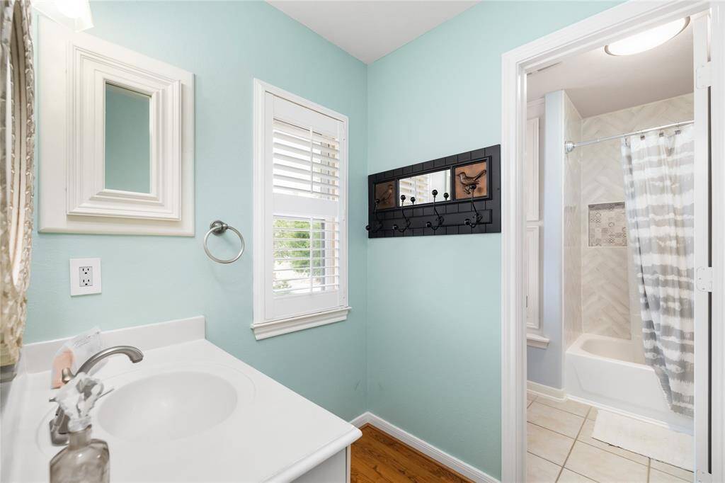 Fresh, bright, and thoughtfully designed. This charming bathroom features soft blue walls, a crisp white vanity with a framed mirror, and a window with plantation shutters that fills the space with natural light. The shower-tub combination is accented with tile details, creating a clean, inviting atmosphere perfect for daily routines or welcoming guests.