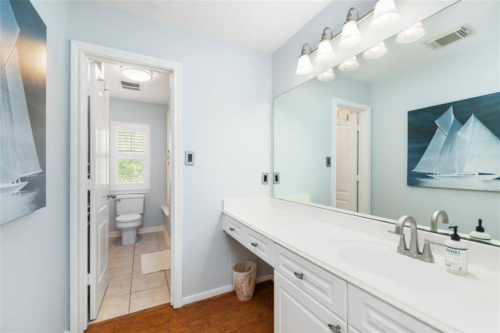A serene and spacious bath designed for everyday ease. This bathroom features a generous vanity with expansive counter space, a wide mirror, and elegant lighting that enhances the room’s airy feel. A separate toilet area benefits from natural light through a nearby window, while the soft blue walls contribute to a calming, spa-like atmosphere.