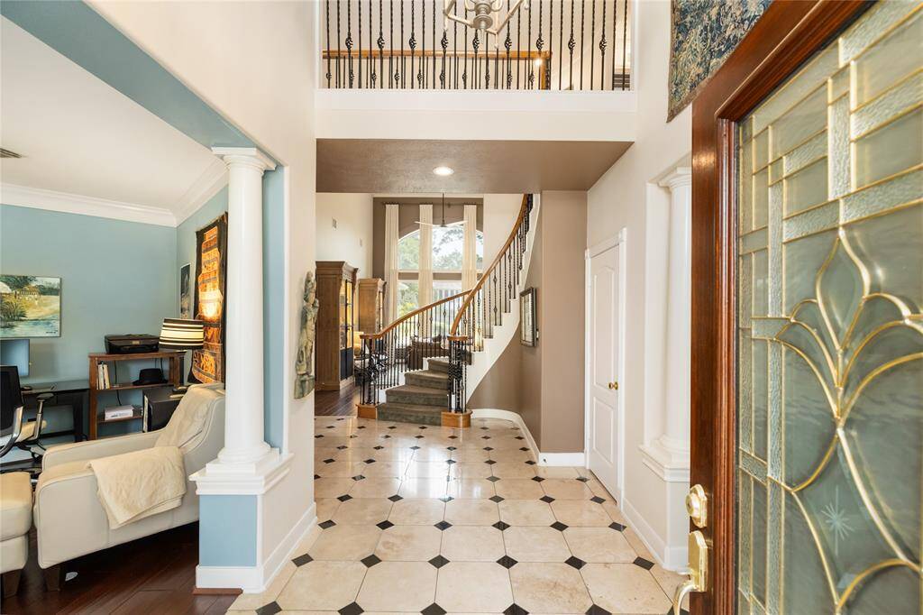 Step into timeless elegance. This grand entryway welcomes you with soaring ceilings, a sweeping staircase with wrought iron accents, and an abundance of natural light. Classic columns frame the open layout, guiding you into a warm living space and a tucked-away office nook—perfect for quiet focus. Patterned tile flooring adds just the right touch of sophistication to this unforgettable first impression.