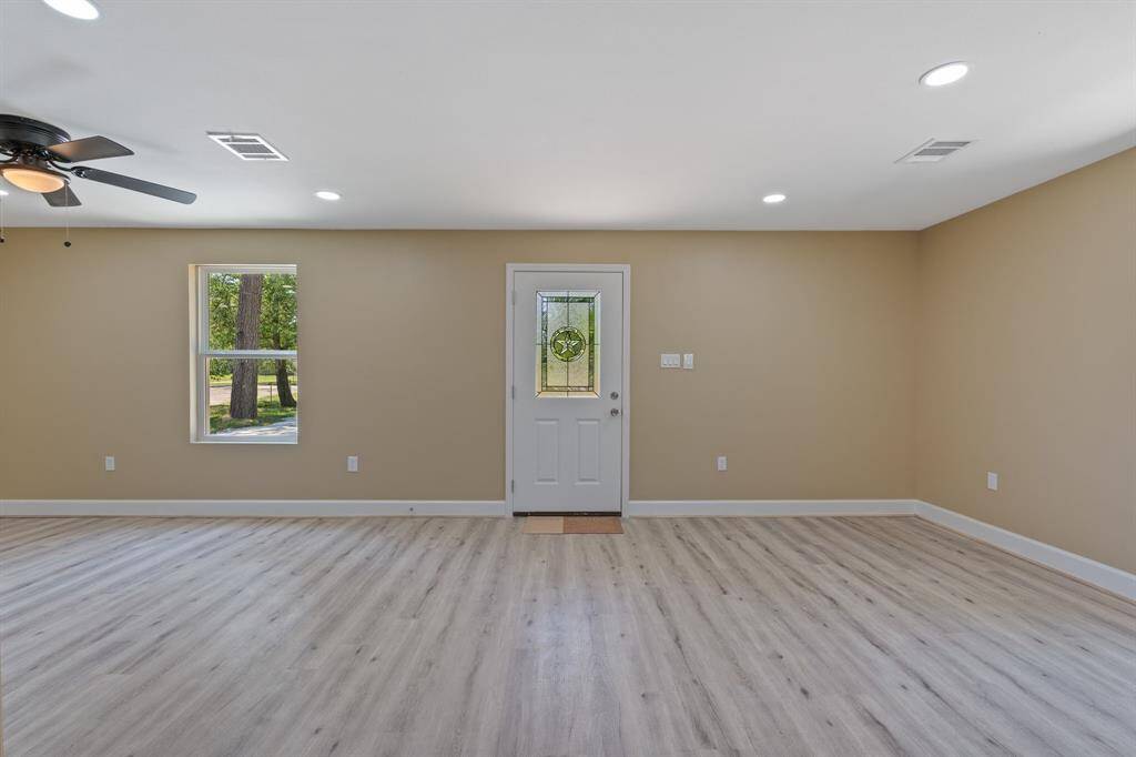 Bright and spacious living room with light wood flooring, neutral walls, and recessed lighting. Features a ceiling fan and a decorative glass front door. Natural light streams in through a side window, offering views of the outdoors.