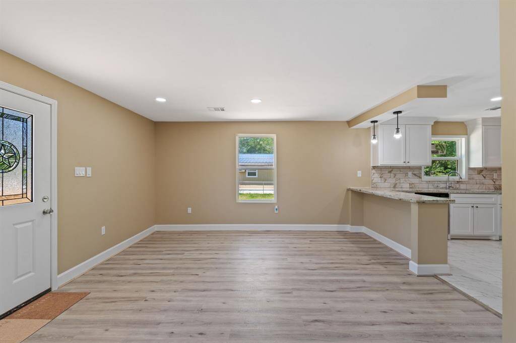 This photo showcases a bright and open living space with light wood flooring and neutral walls. It features a modern kitchen with white cabinets, granite countertops, and pendant lighting. There's a decorative front door and a window providing natural light, creating a welcoming atmosphere.