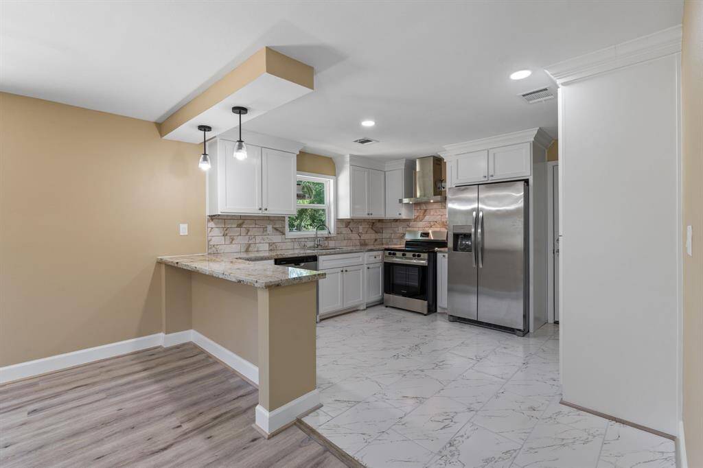 This modern kitchen features white cabinetry, stainless steel appliances, and a stylish tile backsplash. The open layout includes a breakfast bar with pendant lighting, blending seamlessly with the dining area. The combination of wood and tile flooring adds warmth and elegance.