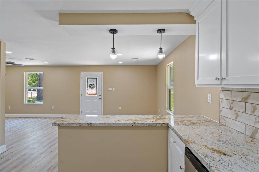 This home features a modern open-concept living area with light wood flooring, a spacious kitchen with granite countertops, white cabinets, and stylish pendant lighting. Natural light flows through large windows, complementing the neutral wall tones.