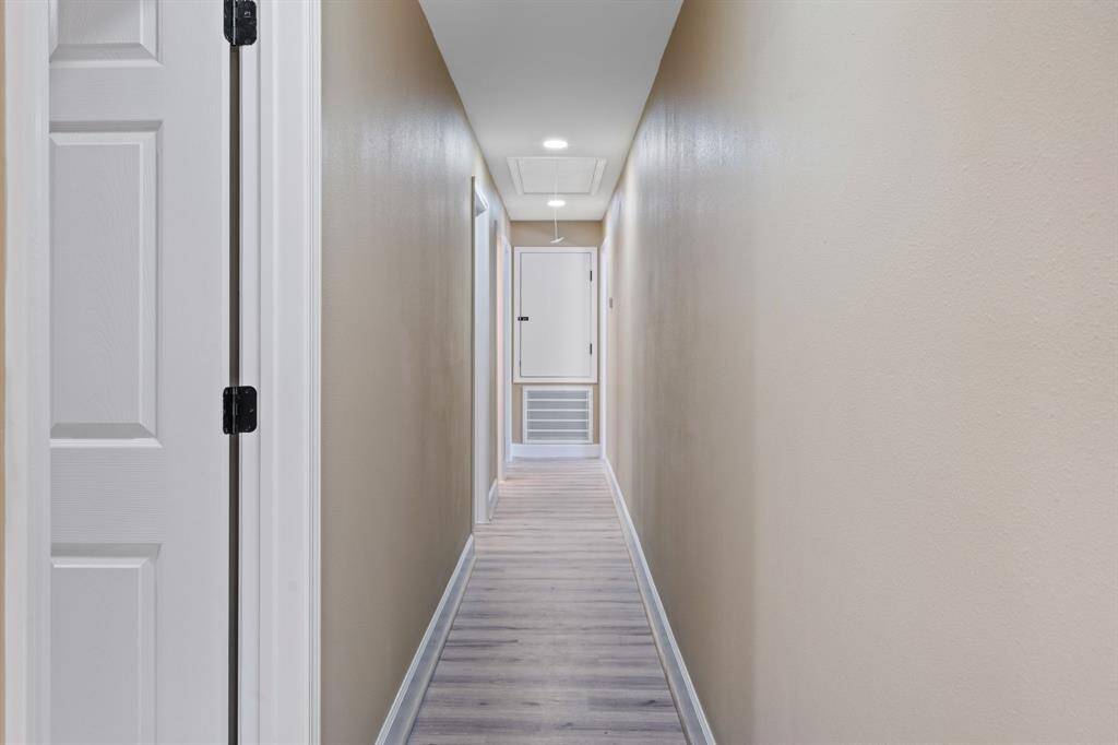 This photo shows a long, narrow hallway with beige walls and light-colored wood flooring. There are white doors on the left and recessed lighting in the ceiling, creating a bright and clean look.