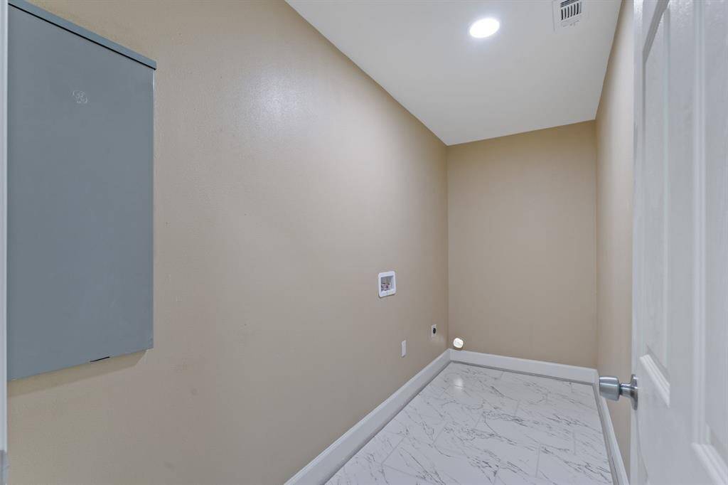 This photo shows a compact utility room with beige walls, a recessed light, and marble-patterned tile flooring. It features hookups for a washer and dryer, with an electrical panel on the wall.