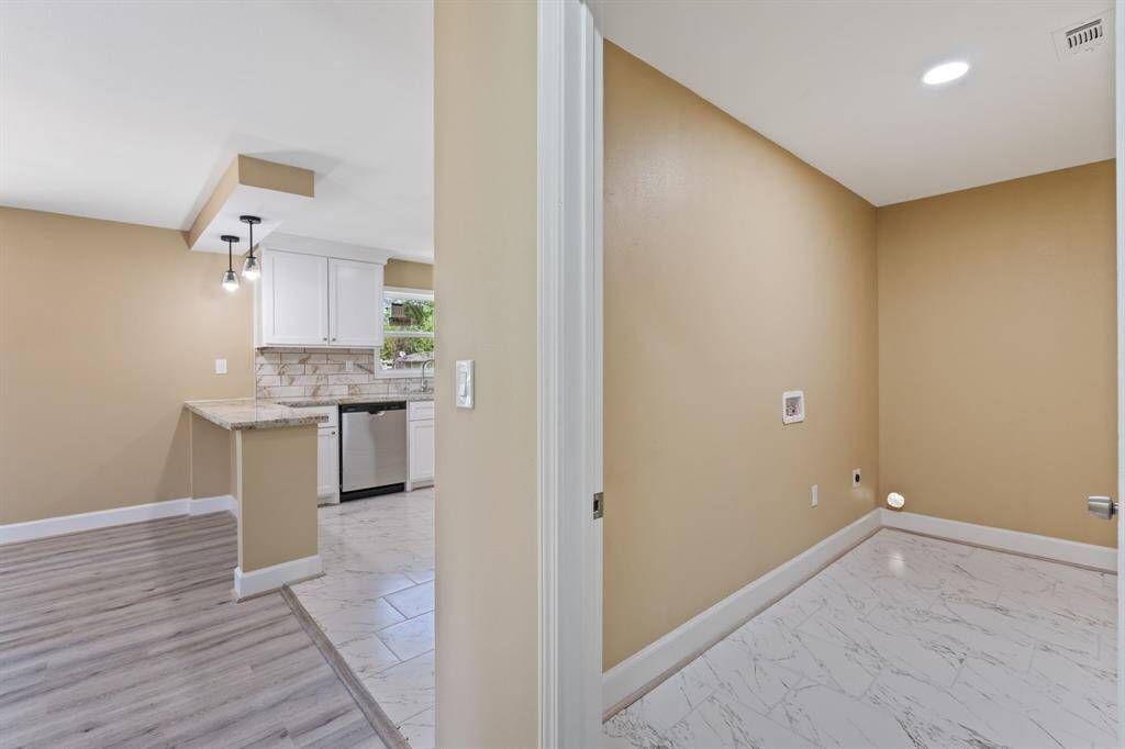 This photo showcases a modern kitchen with white cabinetry, a stone backsplash, and stainless steel appliances. The adjacent room features light-colored walls and tile flooring, suitable for a laundry area. The space is bright and open, with a seamless transition between rooms.