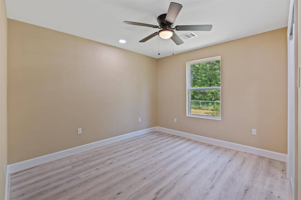 This room features light wood flooring, beige walls, and a ceiling fan. A large window lets in natural light, and there's ample space for furniture.