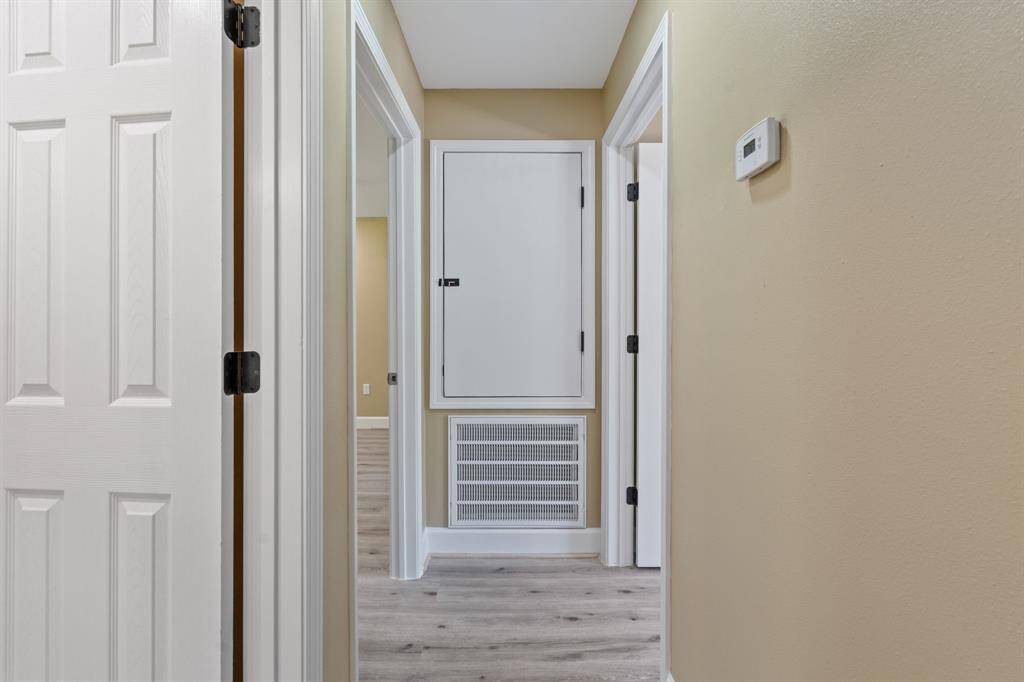 A well-lit hallway with light wood flooring and neutral wall colors. It features multiple white doors, a thermostat on the wall, and an access panel with a vent below.