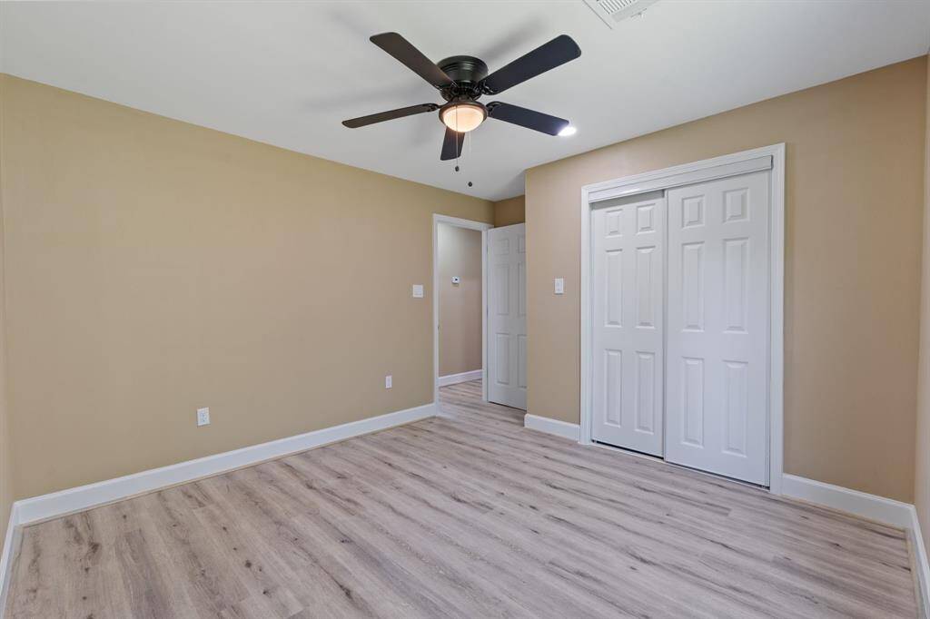 This is a bright bedroom with beige walls, light wood flooring, and a ceiling fan. It features a closet with sliding doors and a doorway leading to another room.