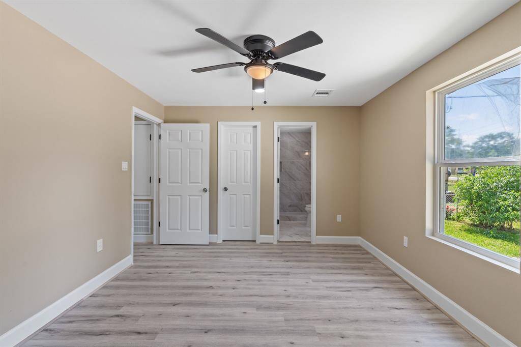 This photo showcases a bright, modern primary bedroom with light wood flooring and beige walls. It features a ceiling fan, large window for natural light, and an en-suite bathroom. The space is airy and inviting, ideal for comfortable living.