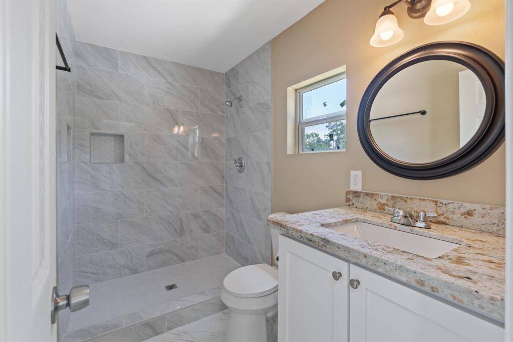 This modern bathroom features a spacious walk-in shower with elegant gray tile, a granite countertop vanity with a stylish round mirror, and ample natural light from a small window.