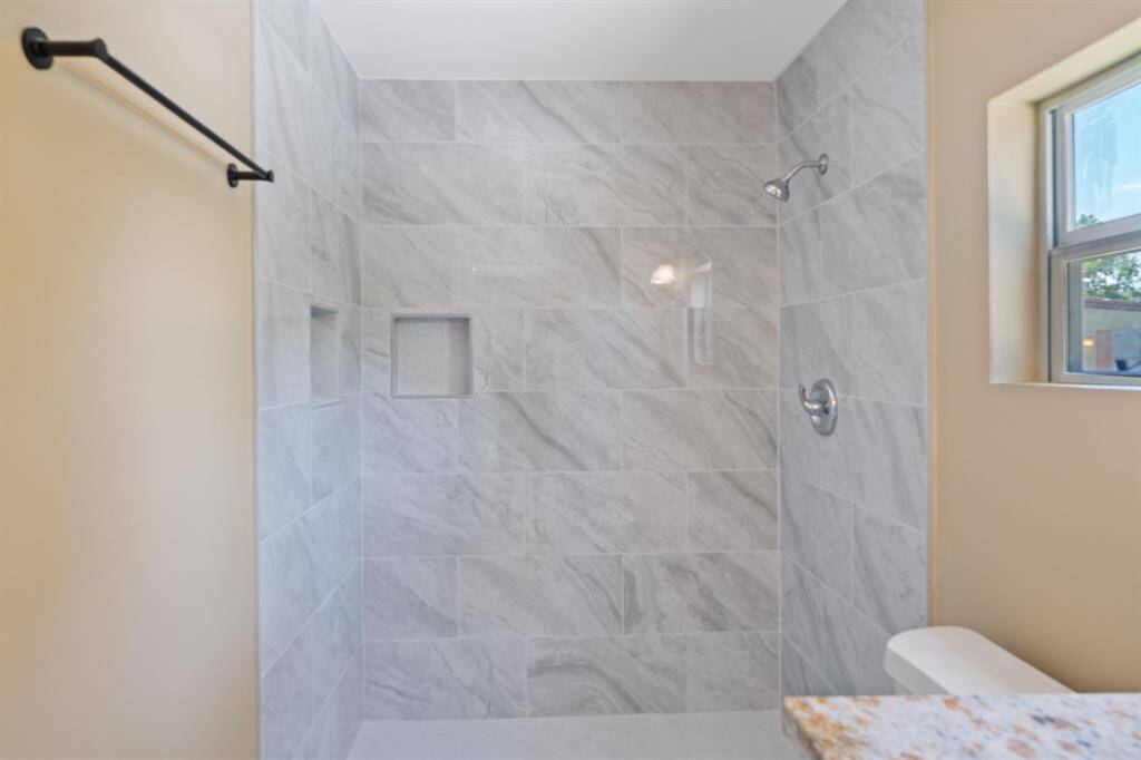 This is a modern bathroom with a spacious, tiled walk-in shower featuring light gray tiles, a built-in shelf, and a window for natural light.
