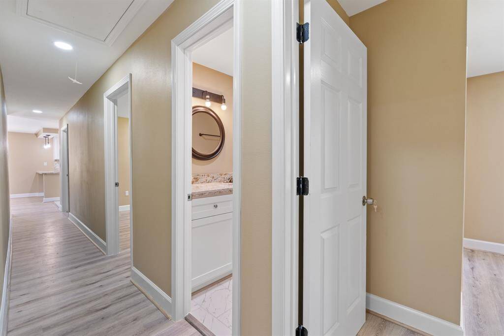 This photo shows a well-lit hallway with light wood flooring leading to multiple rooms. It features a bathroom with a white door, modern fixtures, and a marble countertop. The walls are painted in a warm beige tone, creating a cozy atmosphere.