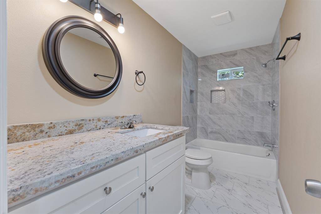 This bathroom features a modern design with a granite countertop, under-mount sink, and sleek fixtures. It has a large, round mirror, tile flooring, and a shower-tub combo with elegant wall tiles. Natural light enters through a small window, creating a bright and inviting space.