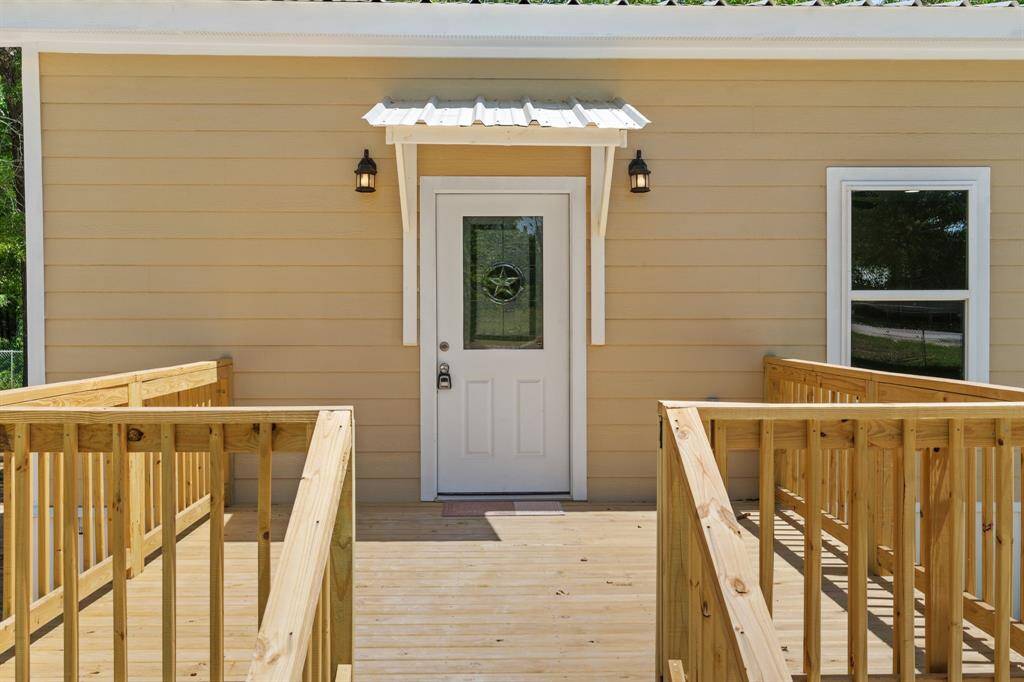 This photo shows the entrance of a home featuring a beige exterior with a white door and decorative glass. The entryway includes a wooden ramp and railing, and is flanked by two outdoor lights.