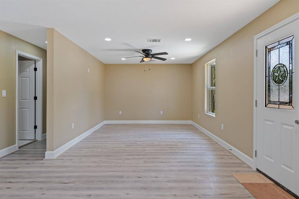 This bright living space features light wood flooring, neutral walls, and recessed lighting. It includes a ceiling fan and a decorative glass front door, offering a modern and inviting atmosphere.