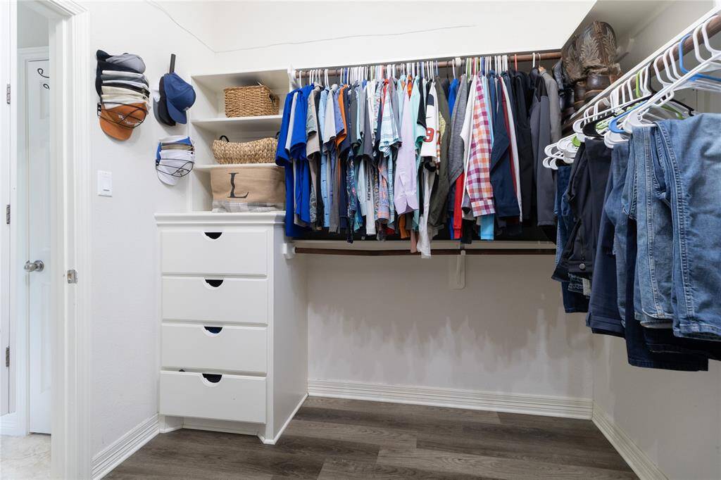 Well-organized walk-in closet with hanging rods for clothing, shelves for storage, and a set of drawers. The space is bright with a clean, modern design and wood-look flooring.