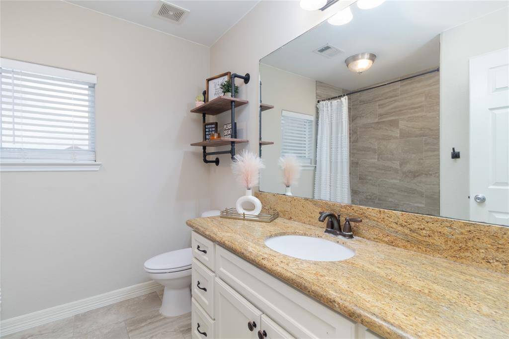 This upstairs bathroom features a spacious granite countertop with an undermount sink, modern fixtures, and ample storage. The neutral tile and light walls create a bright, inviting atmosphere, complemented by a large mirror and open shelving for decor.