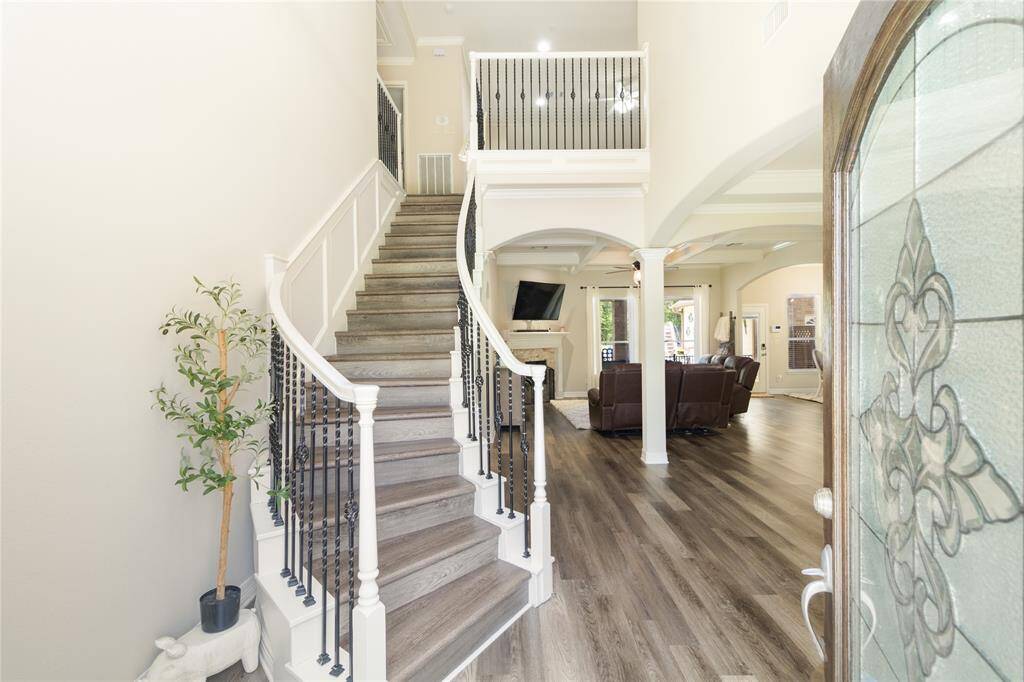This inviting entryway features a stylish curved staircase with wrought iron railings and wood steps, leading up to a second floor with an open railing. The space flows into a bright living area with coffered ceilings and wood flooring, offering a cozy fireplace and modern decor. Natural light fills the room through large windows, enhancing the warm, elegant atmosphere.