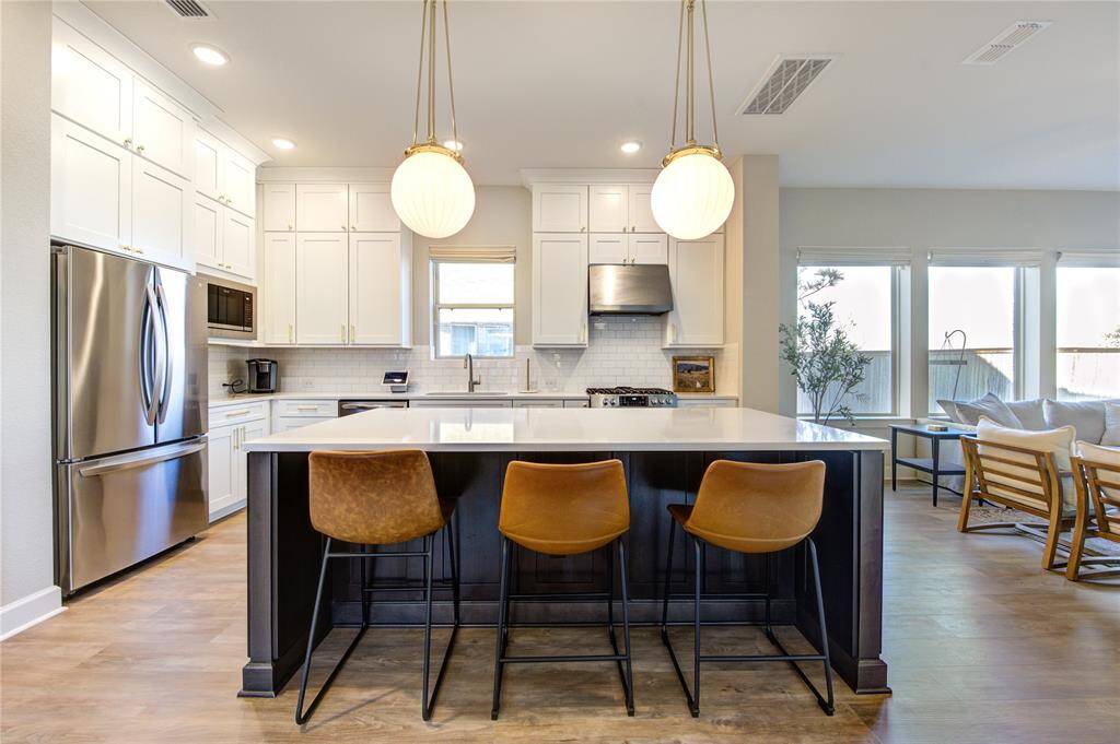 This gourmet island kitchen is a chef’s dream, featuring floor-to-ceiling white cabinetry, quartz countertops, a custom island, a stylish tile backsplash, gorgeous Pendanyt lighting, and stainless steel appliances.