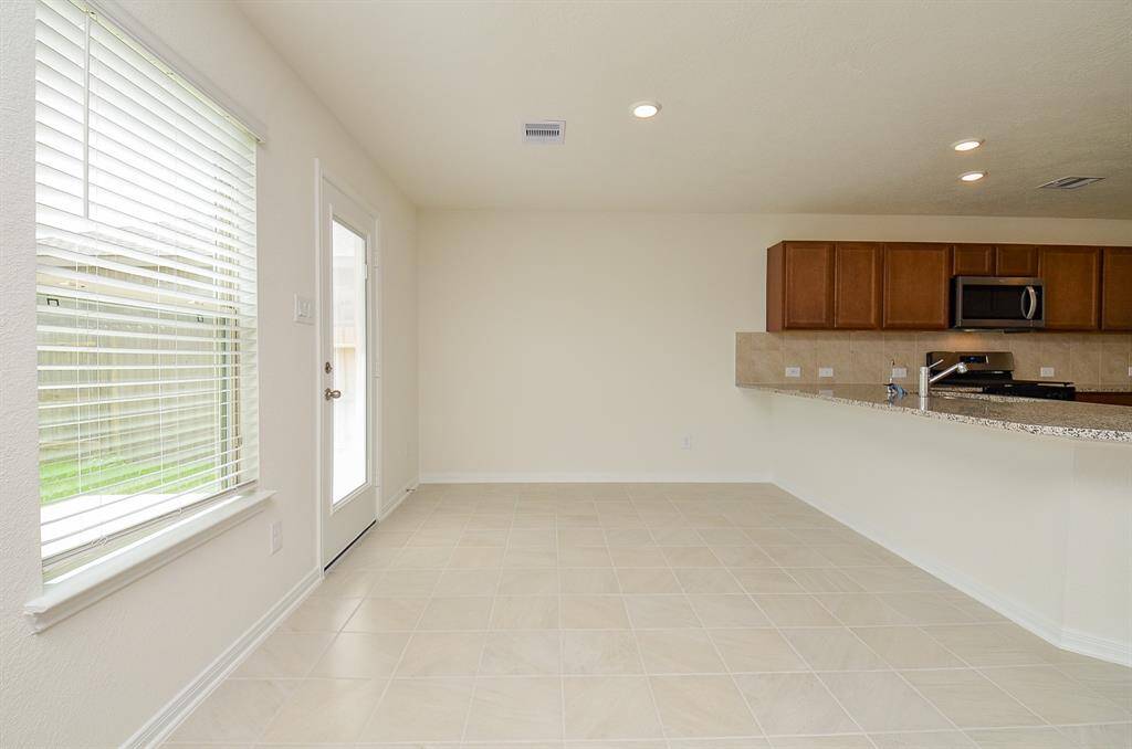 Bright, open kitchen area with clean tile flooring, neutral walls, and abundant natural light from a large window and glass door. Features include modern cabinetry, a granite countertop, and stainless steel appliances, creating a welcoming space for cooking and dining.