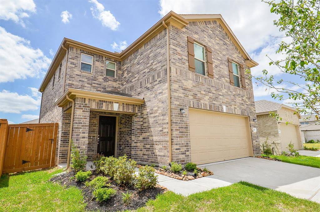 This is a two-story brick home with a two-car garage and a covered entryway. It features a well-maintained front yard with shrubs and a wooden fence. The house has a modern look with large windows and a clean, inviting exterior.