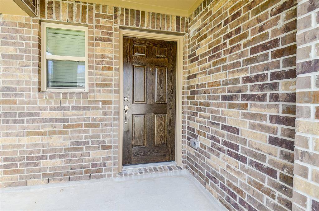 This photo shows a home's front entrance with a solid wood door set in a brick exterior, featuring a small window to the side. The area looks clean and welcoming.