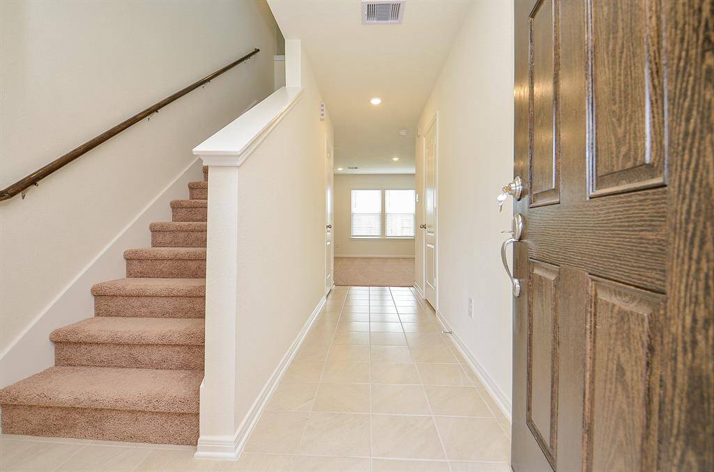 This photo showcases a welcoming entryway with a wooden door, tiled flooring, and a carpeted staircase leading to the upper level. The hallway is well-lit and leads to a bright Living room with large windows.