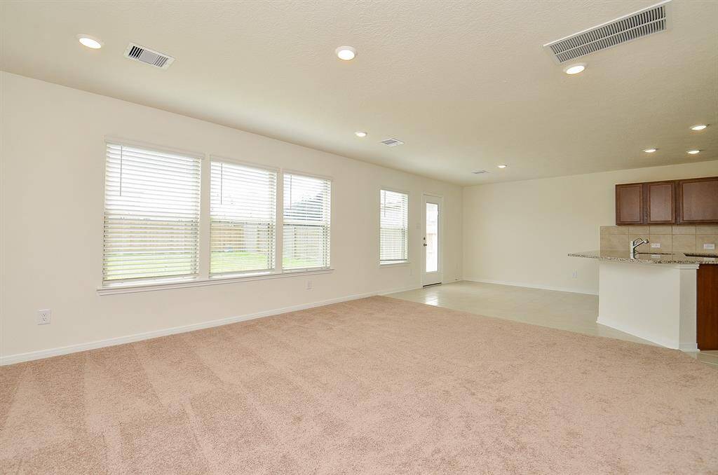 This spacious living area features a blend of carpet and tile flooring, ample natural light from large windows, recessed lighting, and an open layout connecting to the kitchen with granite countertops and wooden cabinets. Ideal for comfortable living and entertaining.