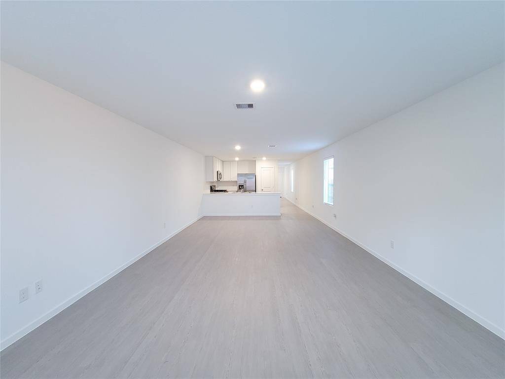 This is a spacious, modern open-plan living area with sleek new floors, leading to a contemporary kitchen with a breakfast bar. The room is well-lit with natural light from windows and recessed lighting, featuring clean, white walls ready for personalization.